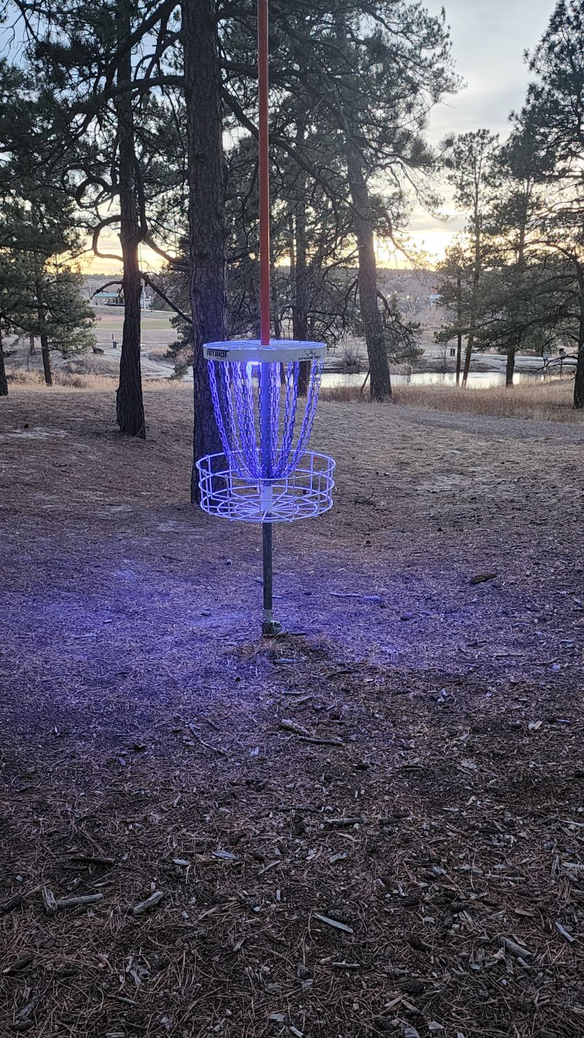 A glowing disc golf basket lit up at dusk on a wooded course
